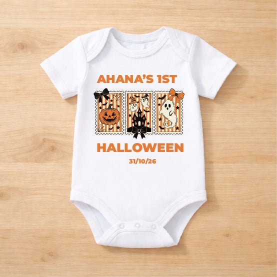 A white short-sleeve baby bodysuit laid flat on a light wood background, featuring an orange and black Halloween-themed graphic with the text 'AHANA'S 1ST HALLOWEEN' and the date '31/10/26'.
