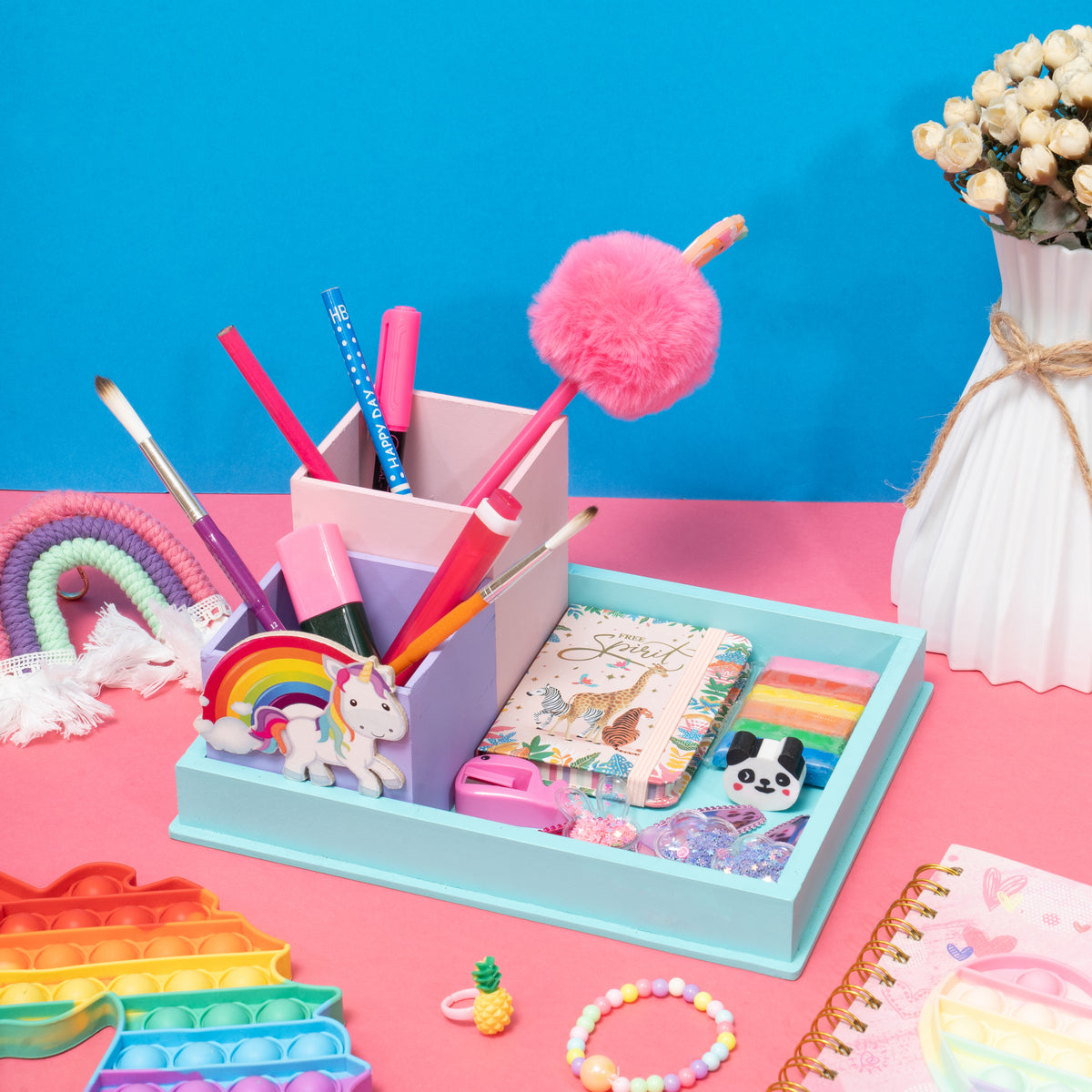 Colorful stationery items including pens, a notebook, and a fidget toy on a pink surface with a blue background.