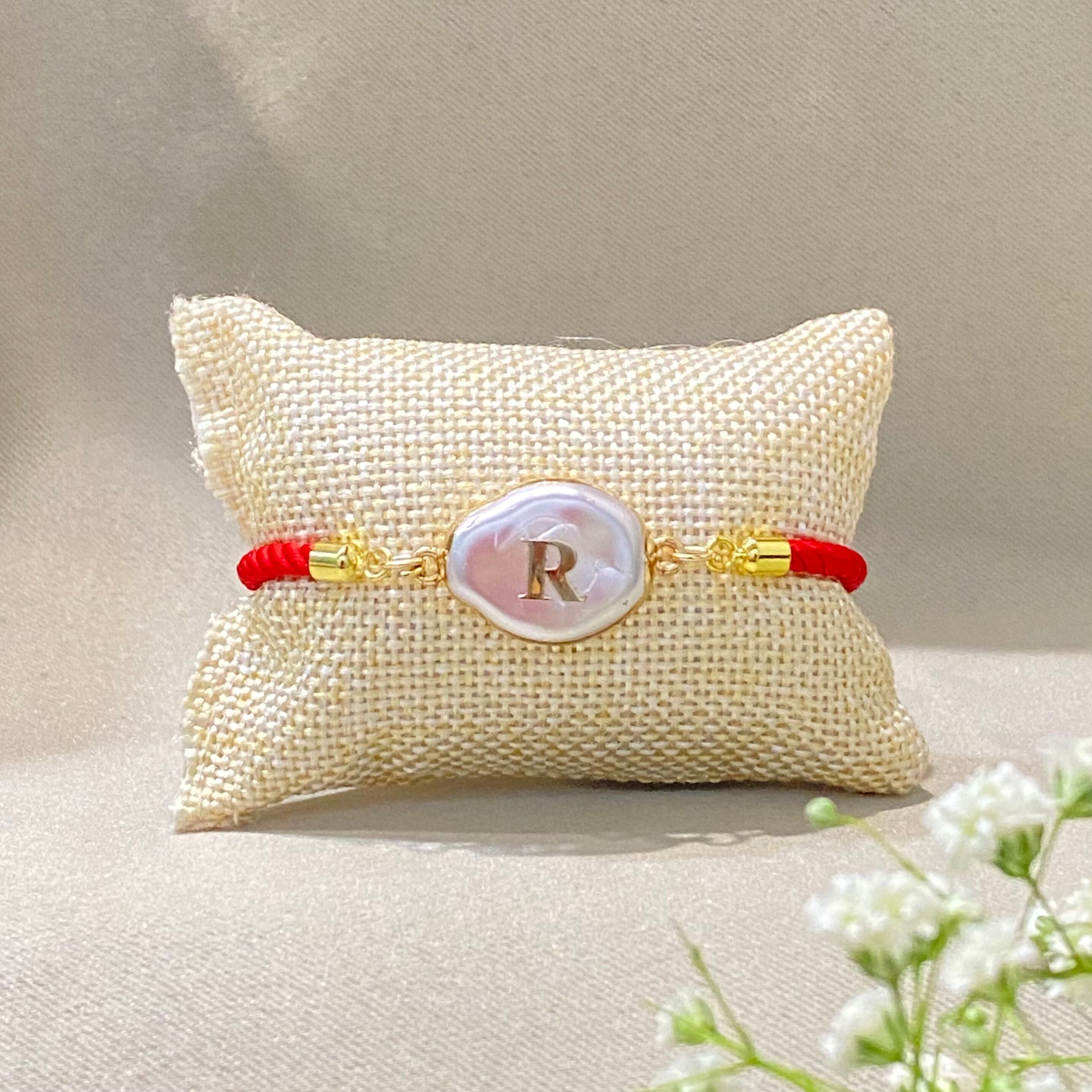 Personalized pearl rakhi with a gold initial charm, natural pearl centre and red adjustable nylon bracelet for Raksha Bandhan.