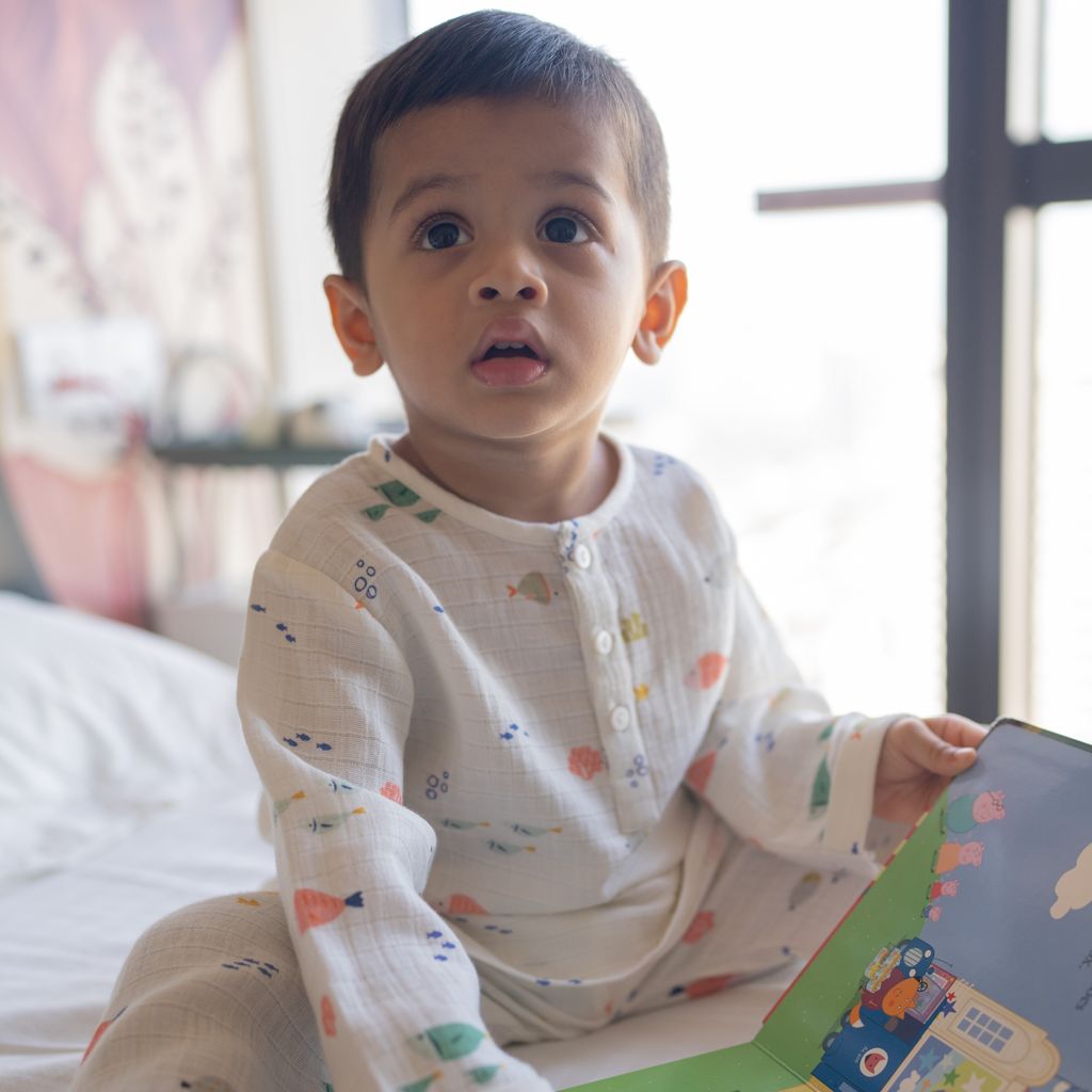 Child in a white outfit with colorful patterns holding a book indoors.