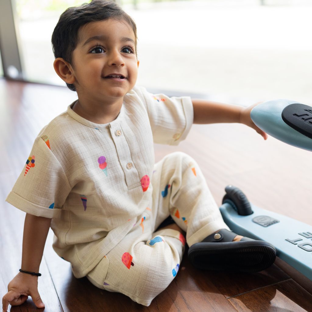 Child sitting on a wooden floor with colorful patterns on a white outfit, holding a blue toy.