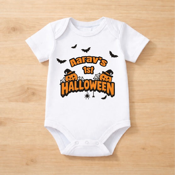 A white short-sleeve baby bodysuit laid flat on a light wood background, featuring a Halloween-themed graphic with the text 'Narav's 1st HALLOWEEN' in orange and black, along with pumpkins, bats, and spiders.