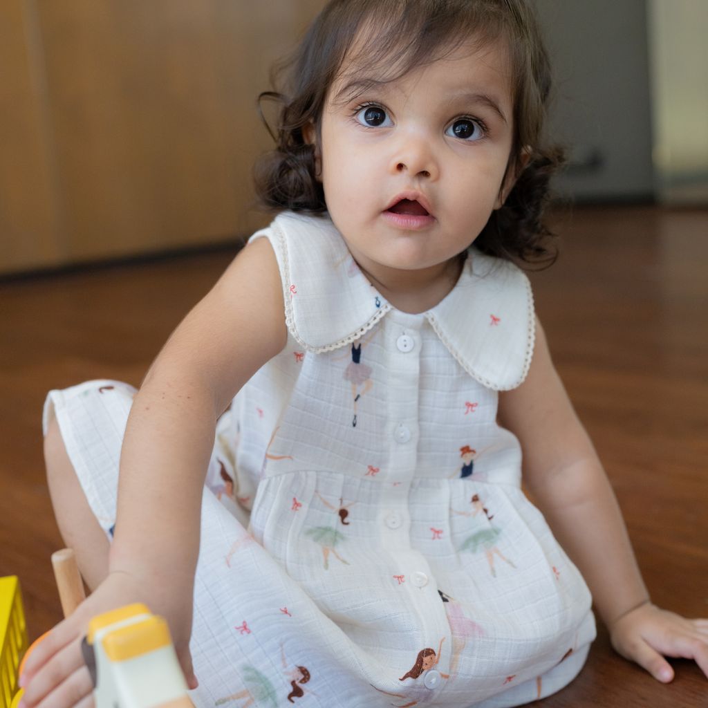 Child in a white dress with a pattern, sitting on the floor holding a toy.