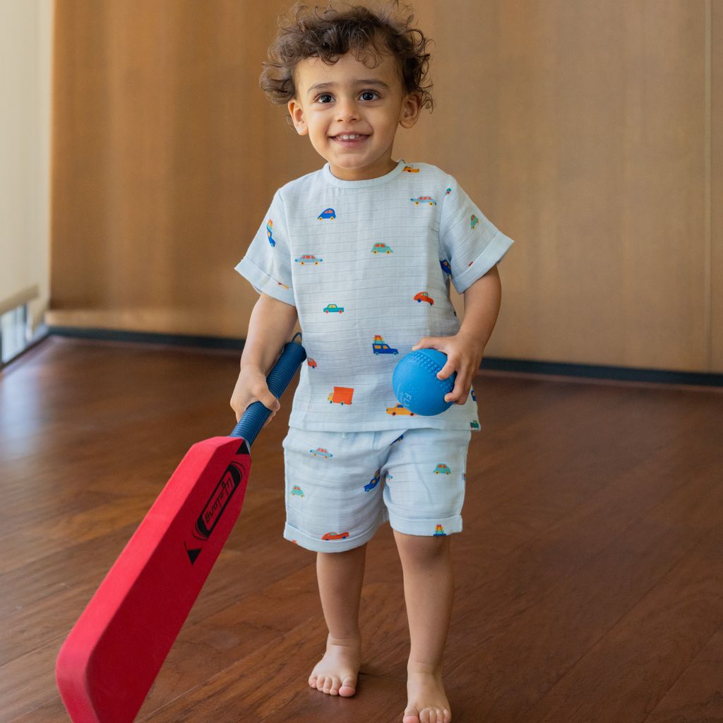 Child holding a red cricket bat and blue ball in a room with wooden floor and wall.