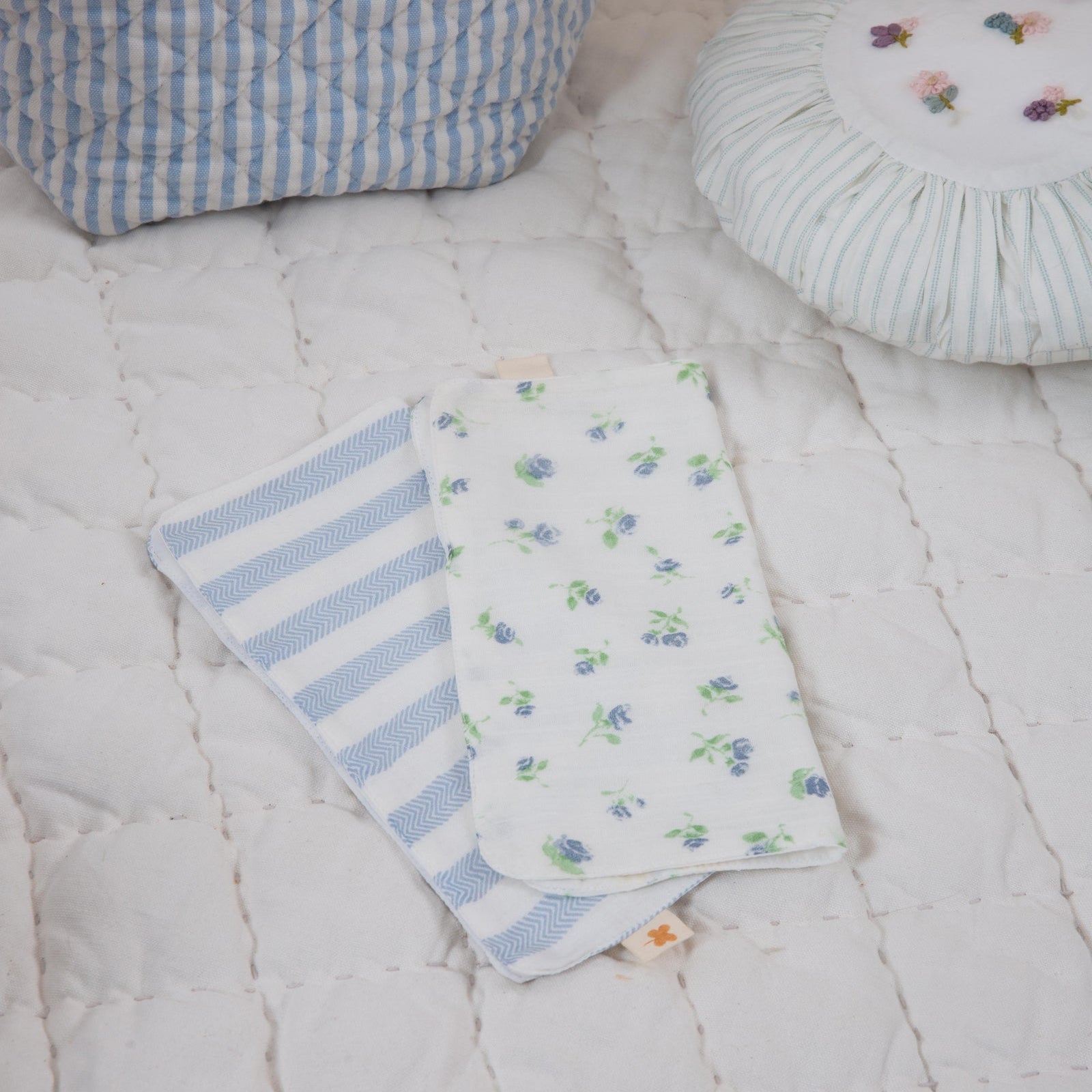 Two baby blankets with blue stripes and floral patterns on a white textured surface, with a basket of stuffed toys in the background.