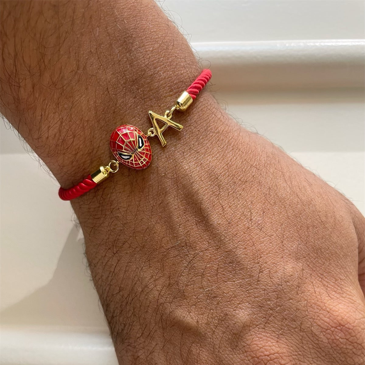 A close-up of a person's wrist and hand wearing a red cord bracelet with gold-tone charms, including a Spider-Man mask and a star-shaped charm.