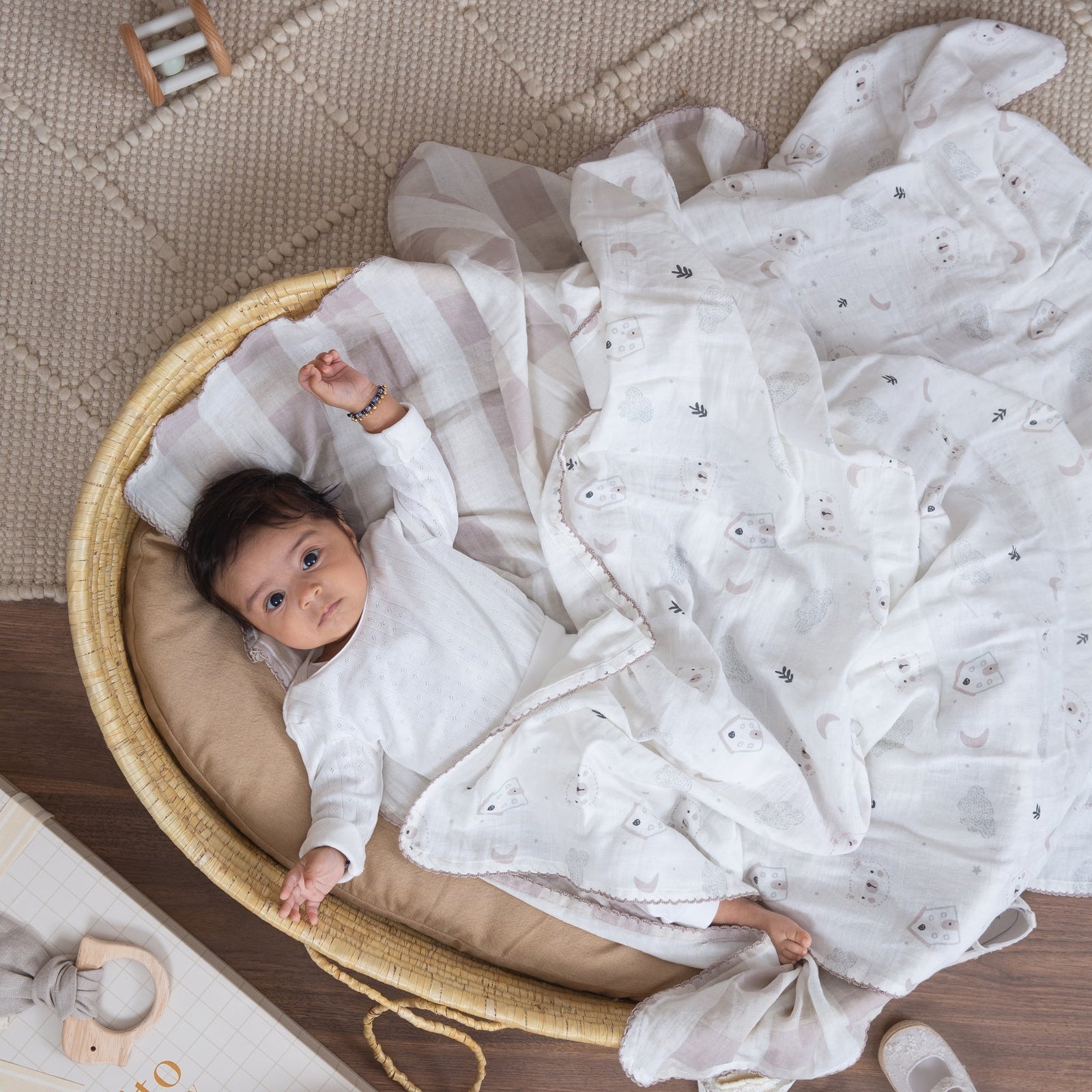 Baby lying in a bassinet with white bedding, surrounded by baby items on a wooden floor.