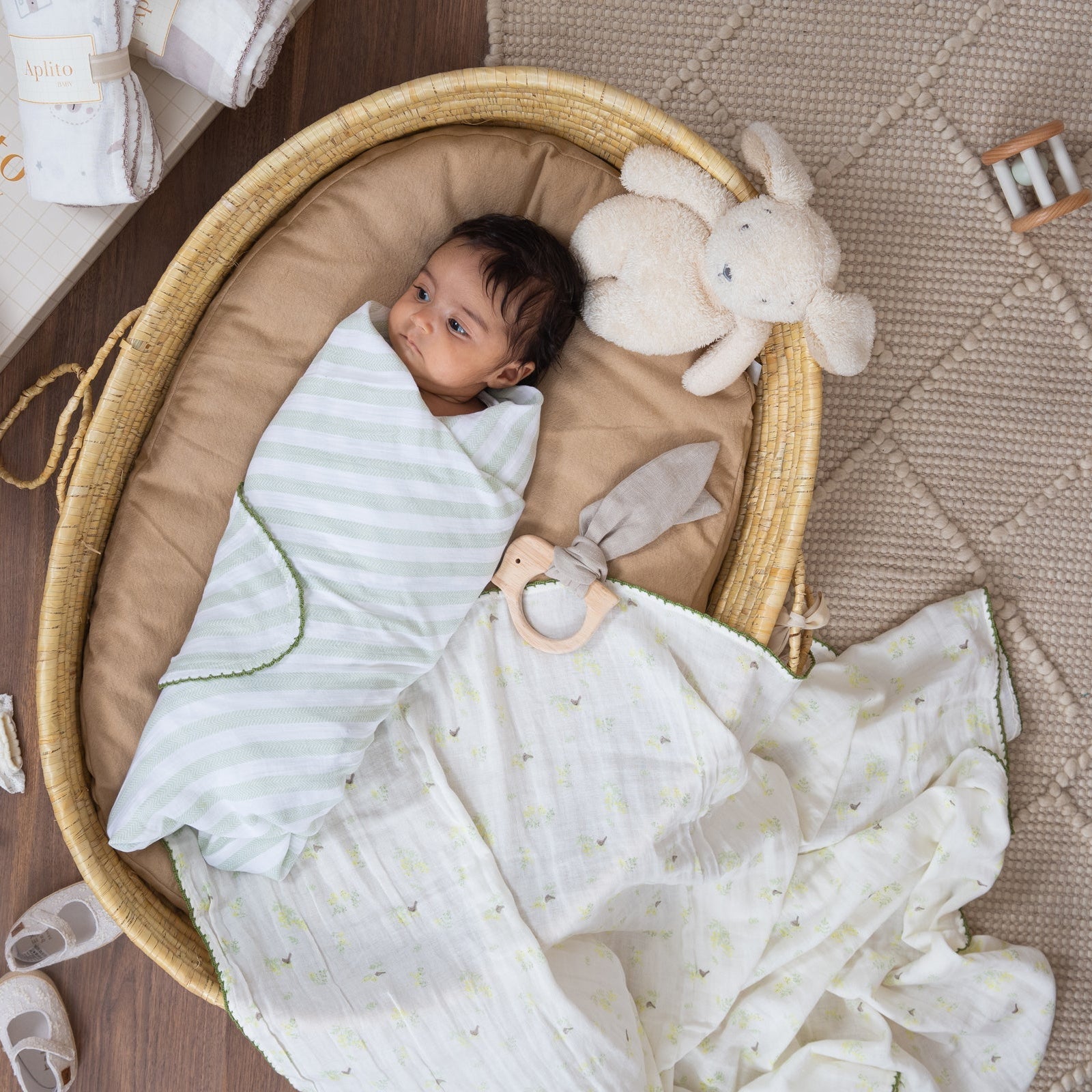 Newborn baby in a bassinet with soft bedding and toys on a wooden floor.