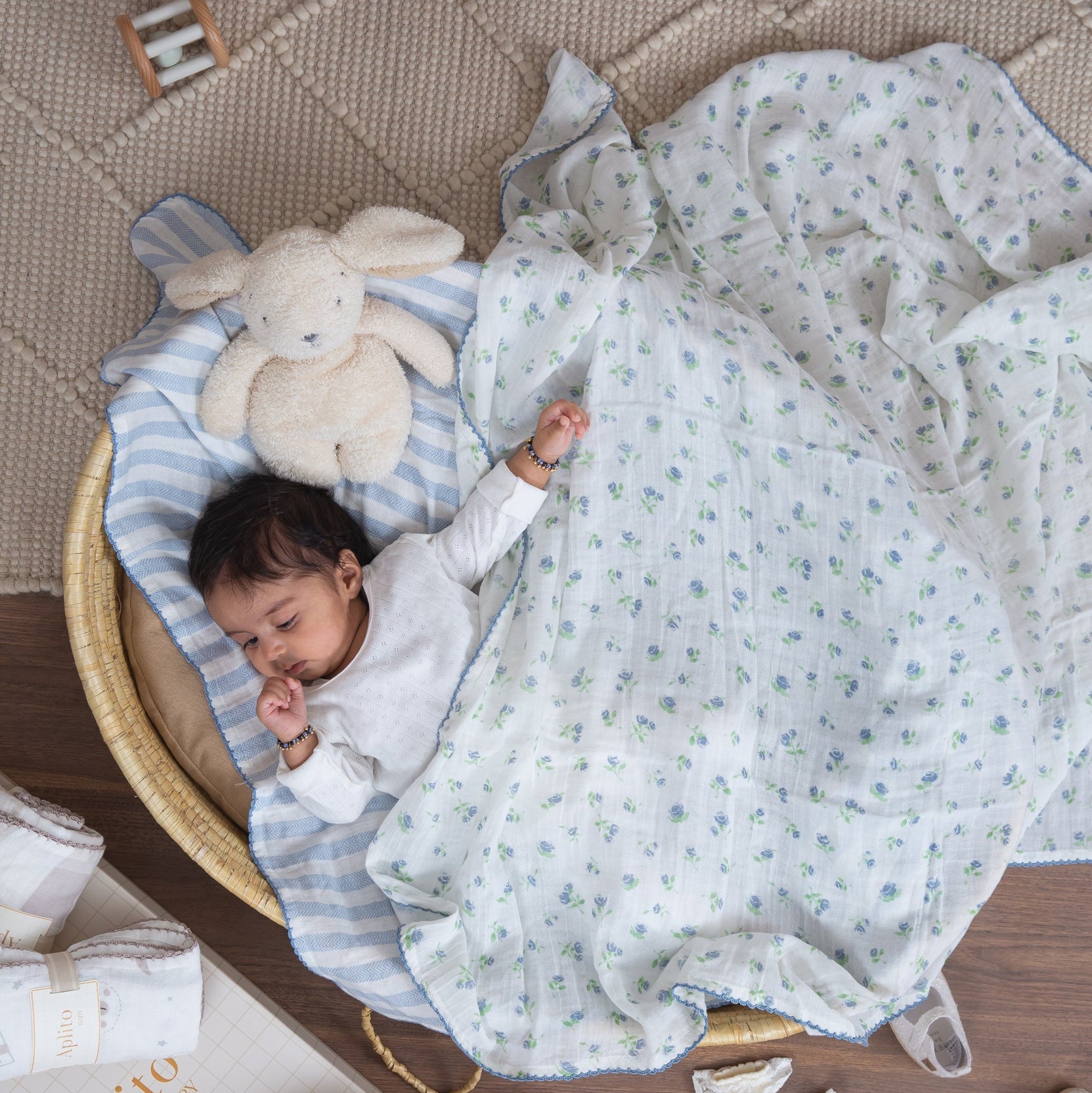 Baby lying in a crib with a white blanket and plush toy, surrounded by books and shoes on a wooden floor.