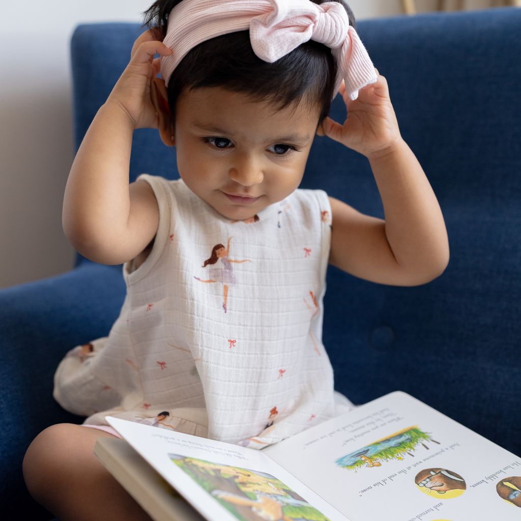 Child sitting on a blue couch with a book open in front of them, wearing a pink headband.