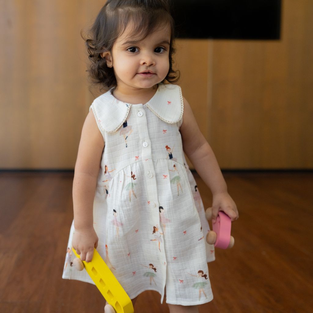 Child in a patterned dress holding a yellow and pink toy on a wooden floor.