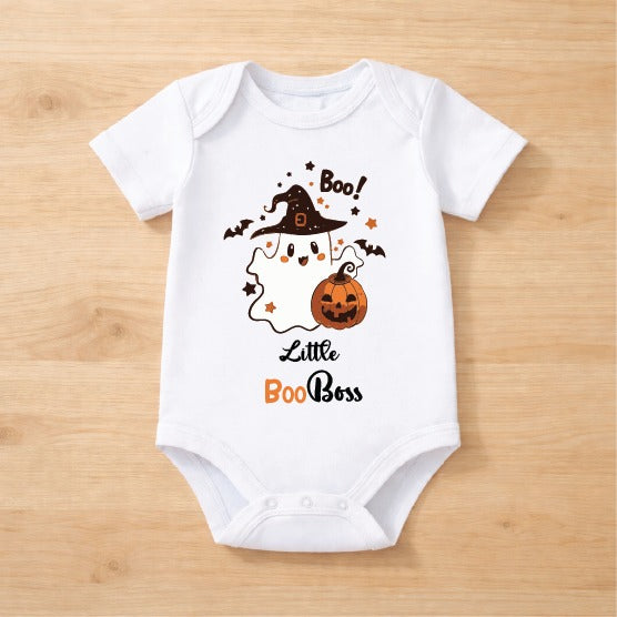 A white short-sleeve baby bodysuit laid flat on a light wood background, featuring a Halloween graphic with a ghost, witch hat, pumpkin, bats, stars, and the text 'Boo! Little Boo Boss'.