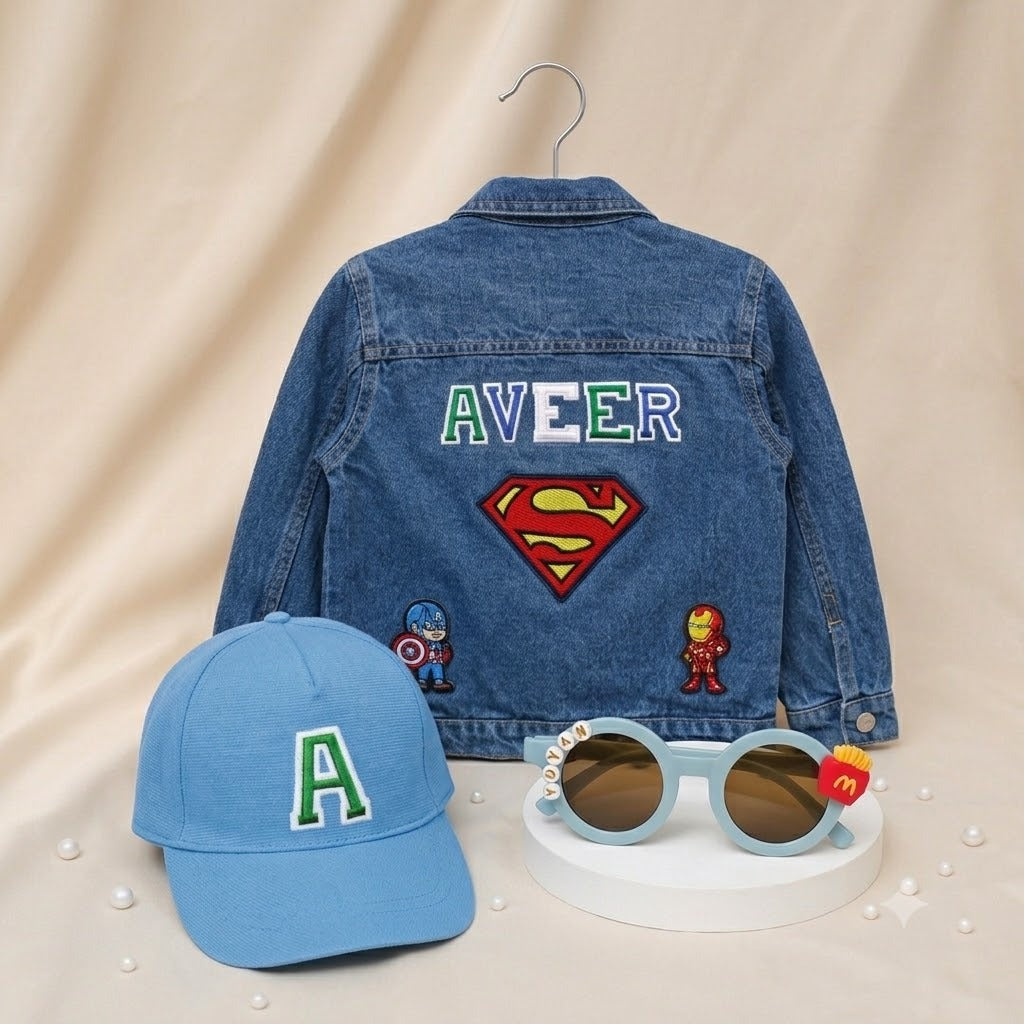 A styled children's outfit display featuring a blue denim jacket with 'AVEER' and a Superman logo on the back, a light blue baseball cap with a green and white letter 'A' on the front, and light blue sunglasses with a small red Superman emblem on the temple, arranged on a beige fabric backdrop.
