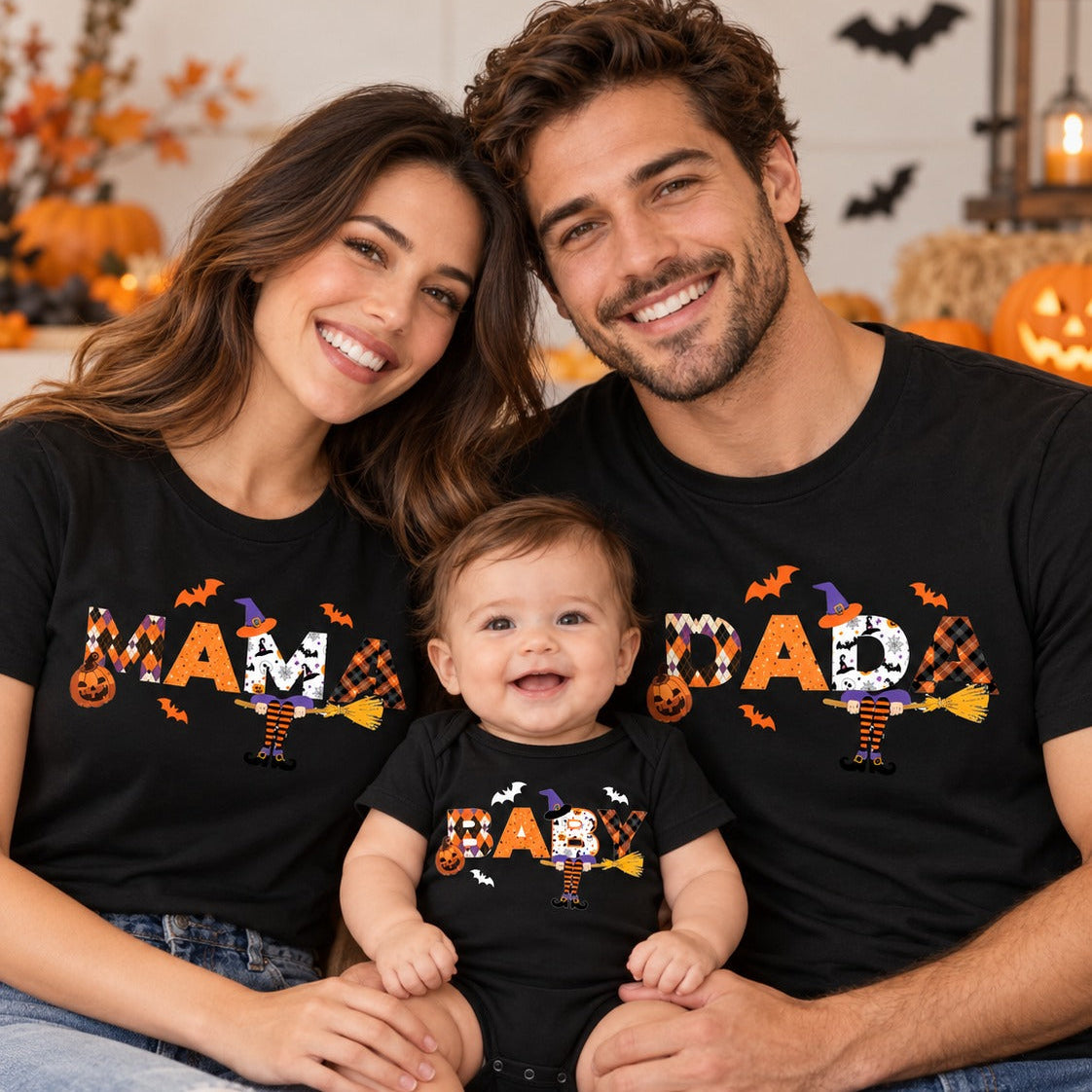 A smiling family of three wearing matching black graphic T-shirts and jeans, posed together in a cozy Halloween-themed room. The woman wears a shirt reading 'MAM' with a witch and pumpkin design, the man wears a shirt reading 'DAD' with a spider and bat design, and the baby wears a matching shirt reading 'BABY' with a cute character.