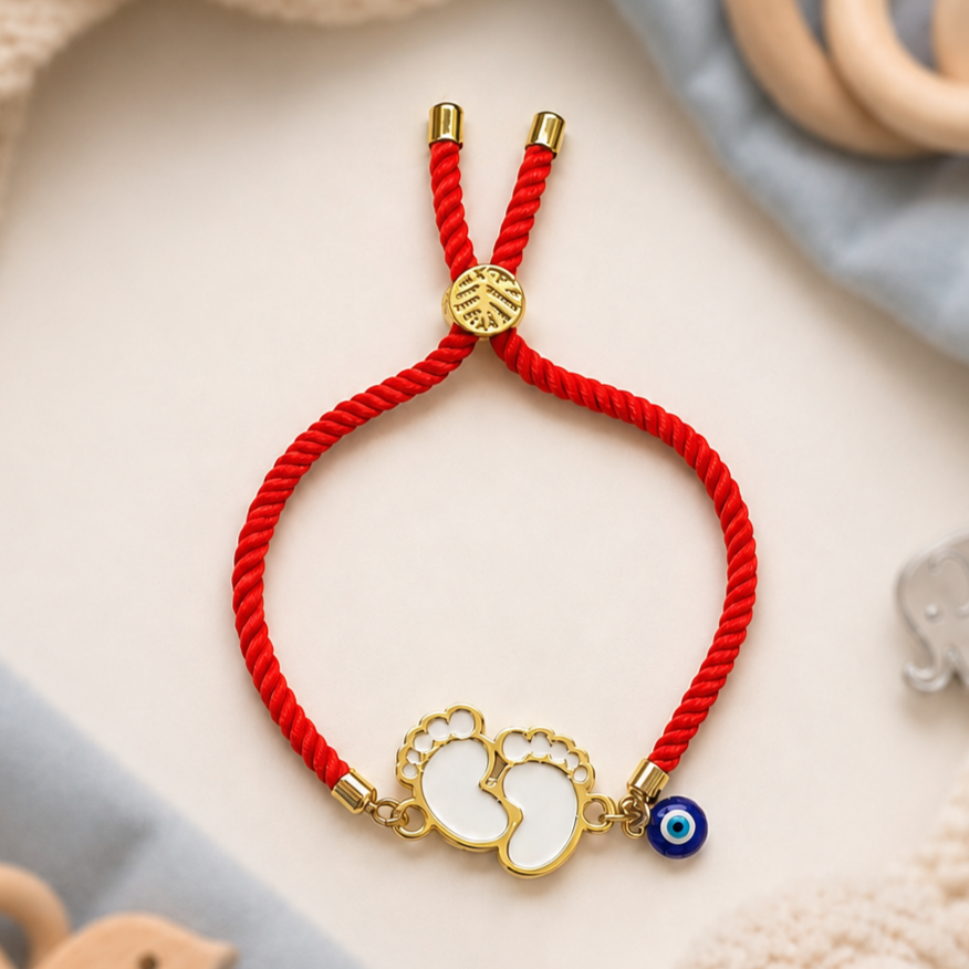 Newborn baby feet rakhi with white acrylic foot charms, gold detailing, evil eye charm and adjustable red nylon bracelet.