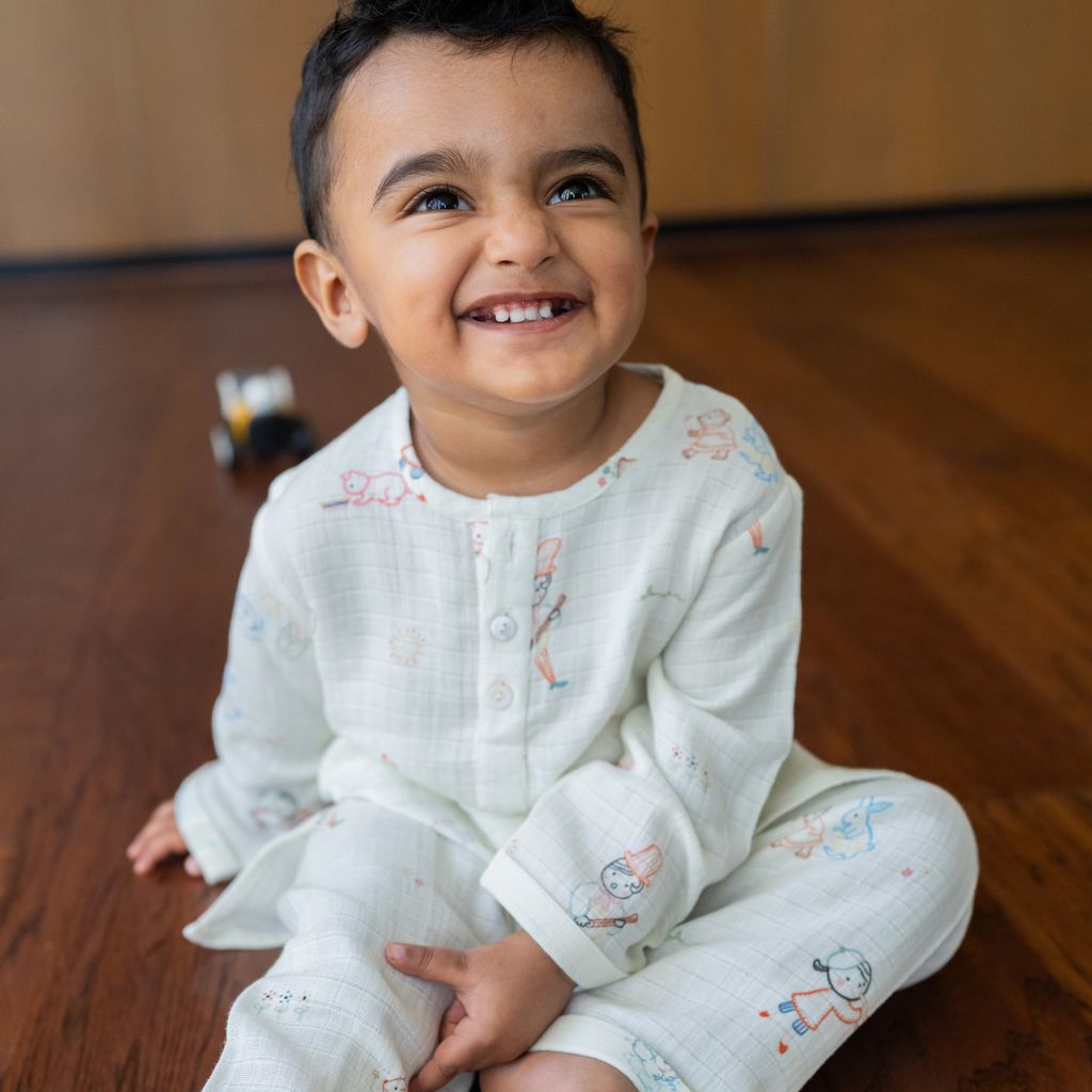 Child wearing a white outfit with colorful patterns sitting on a wooden floor.
