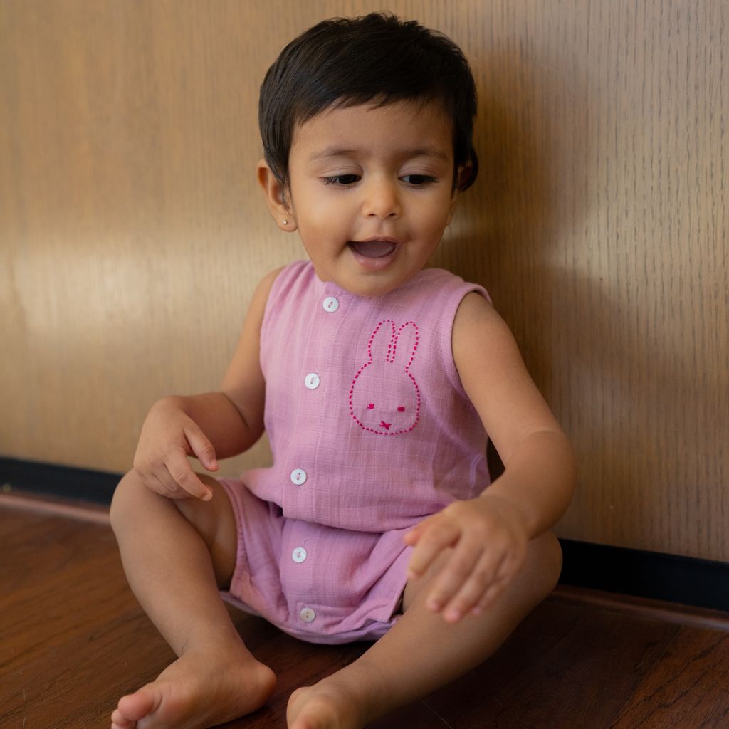Child wearing a pink outfit with a bunny design sitting on the floor against a wooden wall.