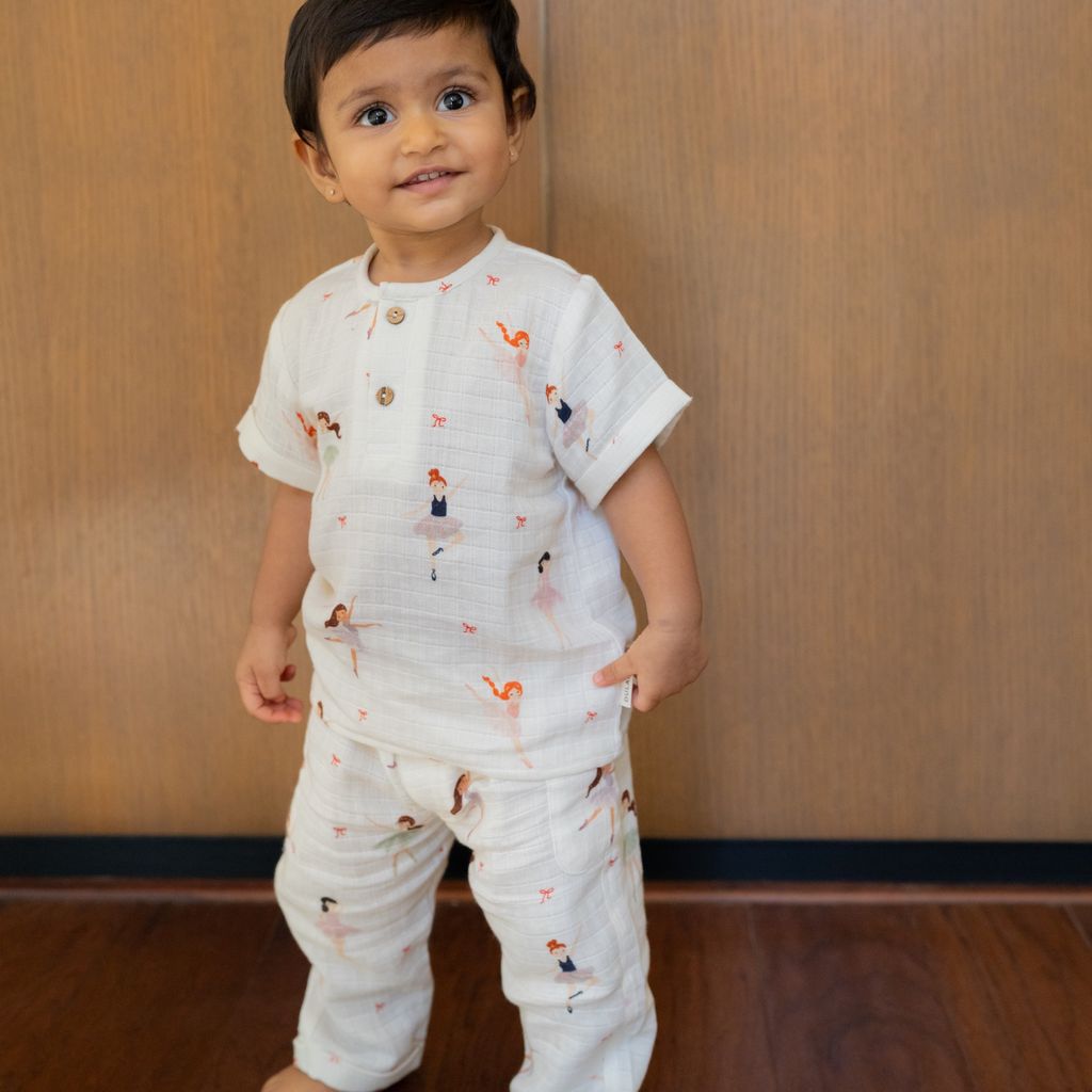 Toddler wearing a soft white muslin co-ord set featuring a short-sleeve shirt and jogger pants with delicate ballerina print details and wooden buttons, standing indoors against a wooden backdrop.