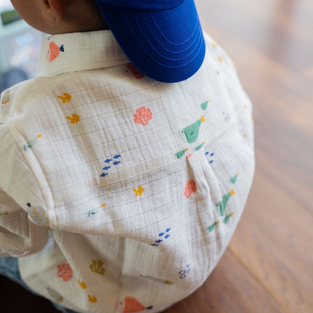 White long-sleeve children's button-down shirt featuring an all-over colorful fish and underwater print, with a classic collar, front button closure, and chest pocket, displayed on a light background.